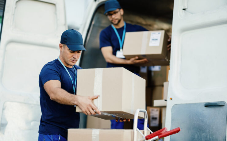 Young courier and his colleague unloading cardboard boxes from delivery van.